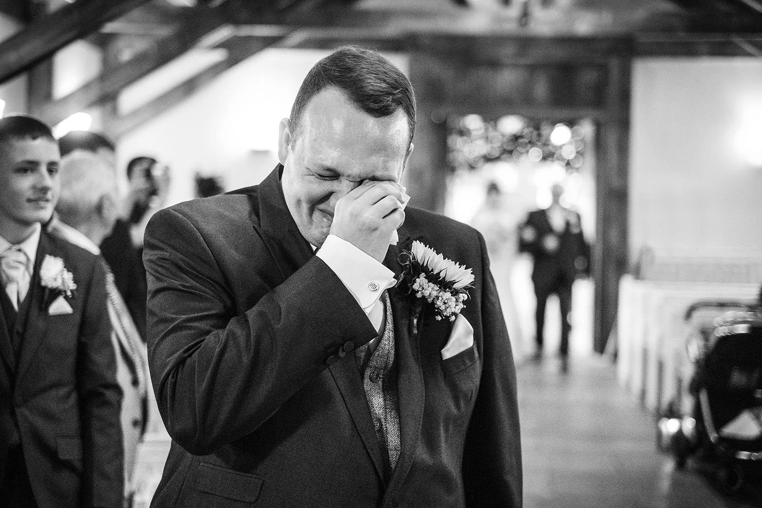 South Causey Inn wedding: groom wiping away tears with his hand as the bride walks up the aisle behind him, black and white