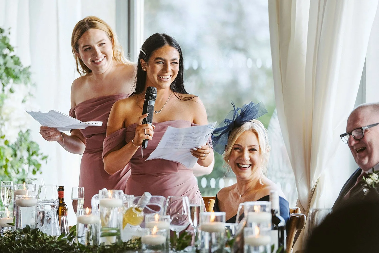 Newton Hall wedding: bridesmaid in a dusky pink dress reading a speech into a microphone at the top table