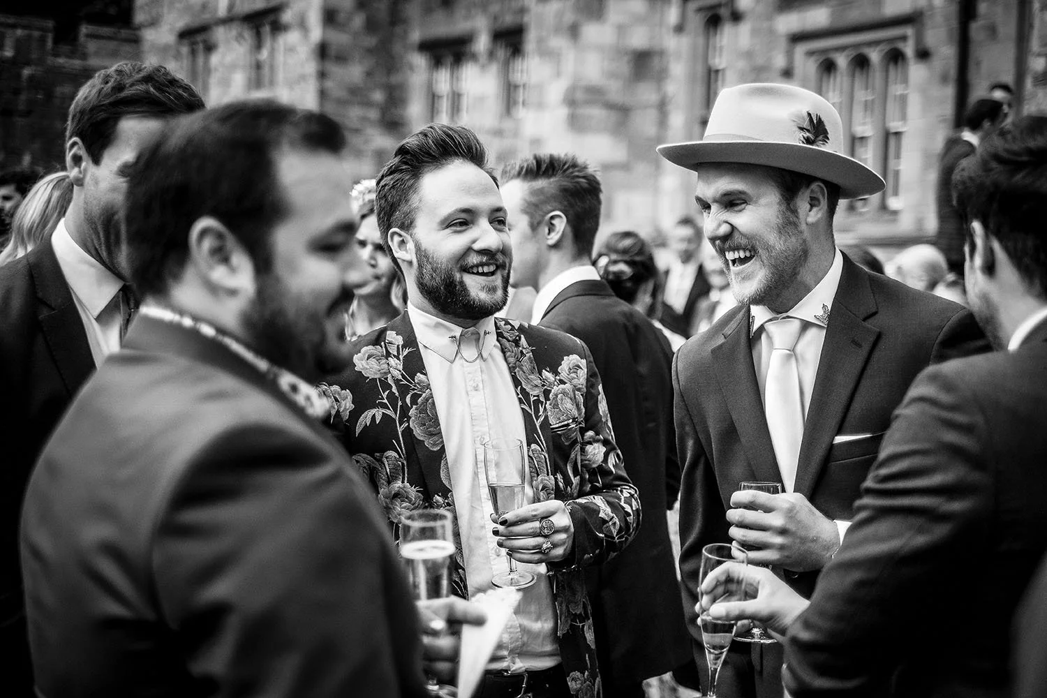 Black-and-white wedding reception photo of well-dressed guests laughing and holding champagne glasses outdoors