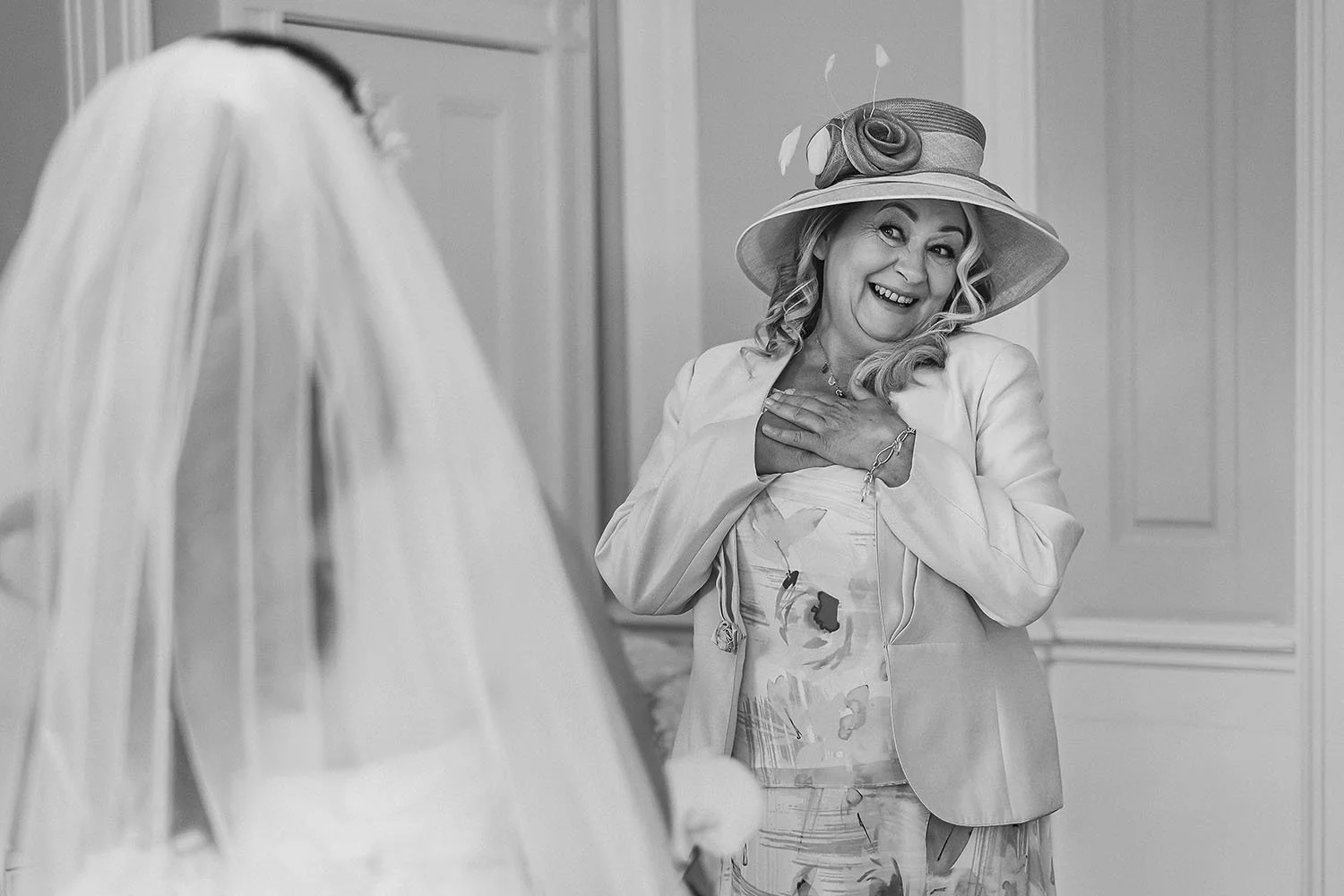 Beamish Hall wedding: mother of the bride in a wide-brimmed hat with her hand on her heart as she sees the bride in her veil