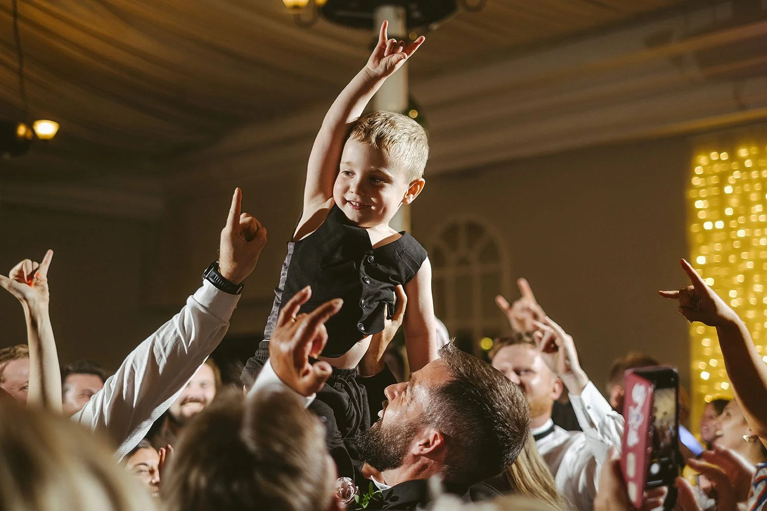 Beamish Hall wedding: young boy on a man's shoulders pointing upwards as guests dance with their arms raised.