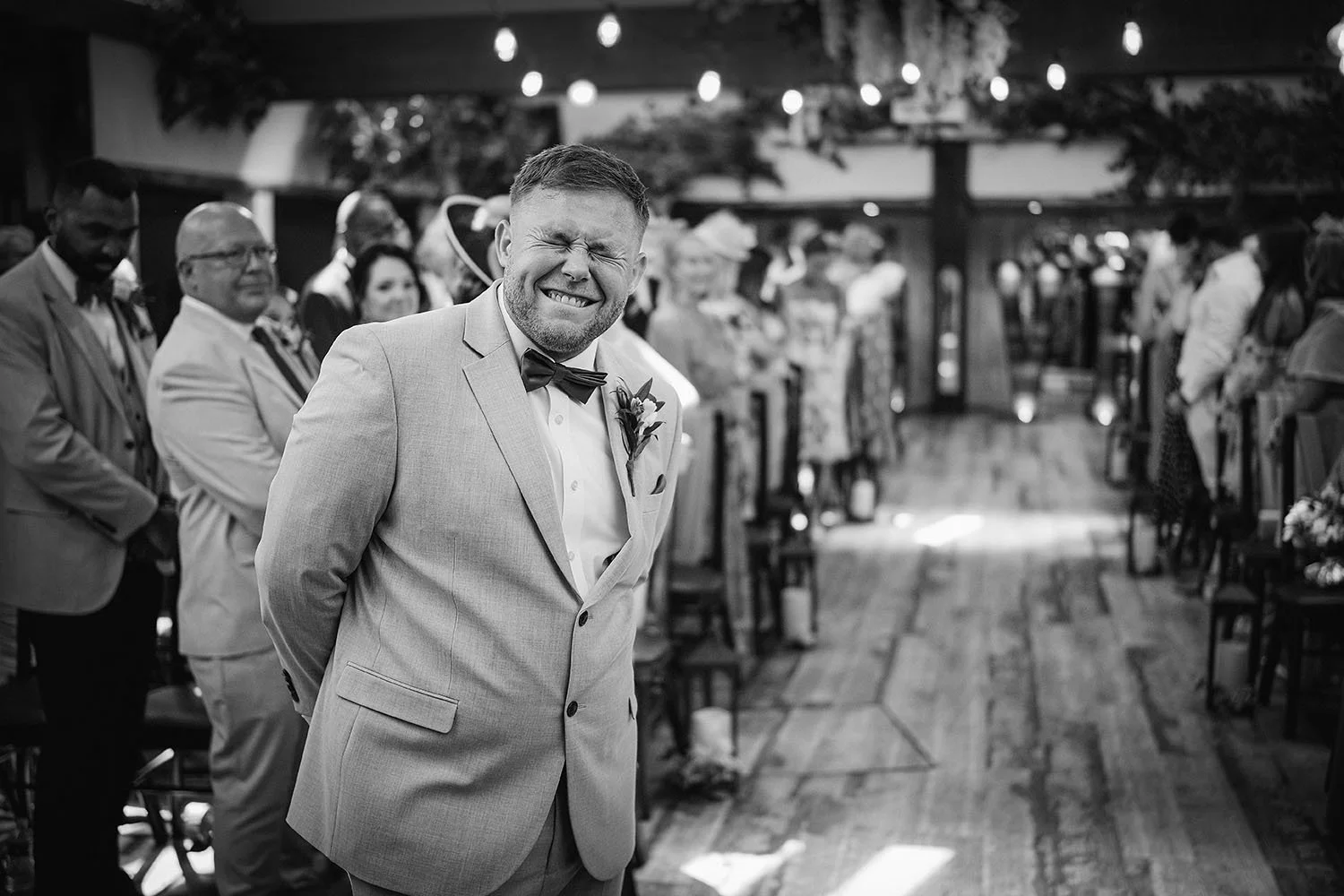 South Causey Inn wedding: groom in a bow tie laughing with his eyes shut as he waits at the aisle in a barn, in black and white.