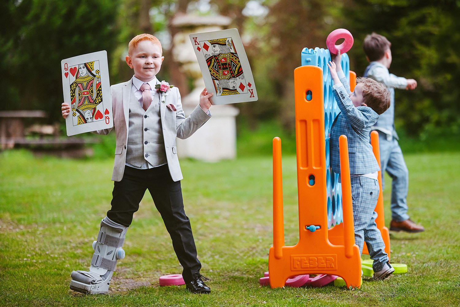 Bowburn Hall wedding: young boy in a suit holding giant playing cards on a lawn, his foot in a walking boot