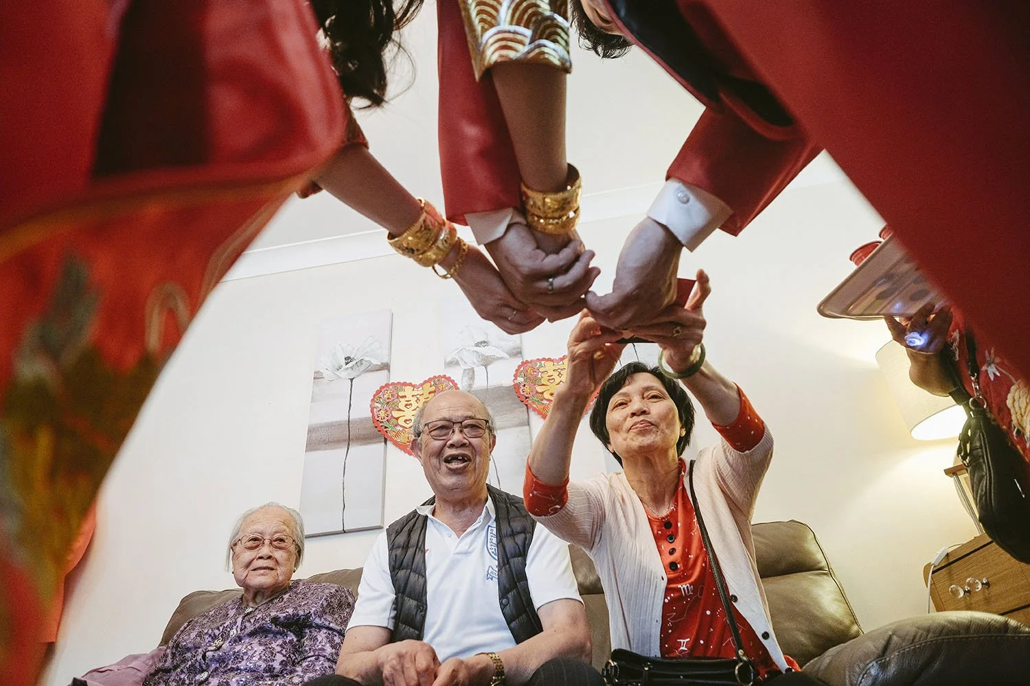 True Jesus Church wedding: grandparents laughing on a sofa as hands in red sleeves with gold bangles reach in, seen from below.