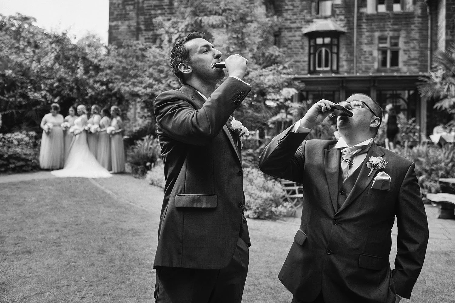 Two men drinking shots on a lawn while bridesmaids and the bride gather in the distance, in black and white.