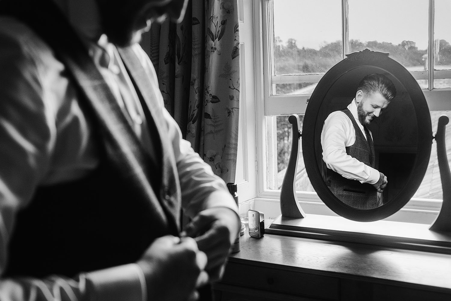 Wynyard Hall wedding: groom reflected in an oval mirror on a windowsill as he fastens his cuffs, black and white