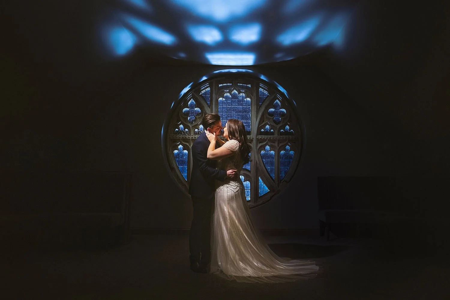 Ellingham Hall wedding: bride and groom kissing in front of a round stained glass window in moody blue light.