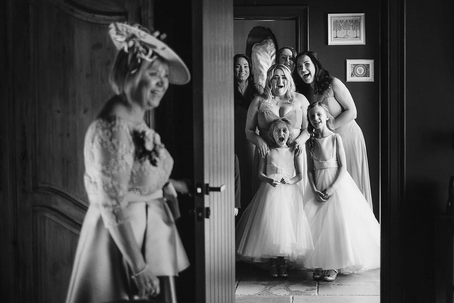 Mother of the bride smiling as bridesmaids and flower girls peek around a door, in black and white.
