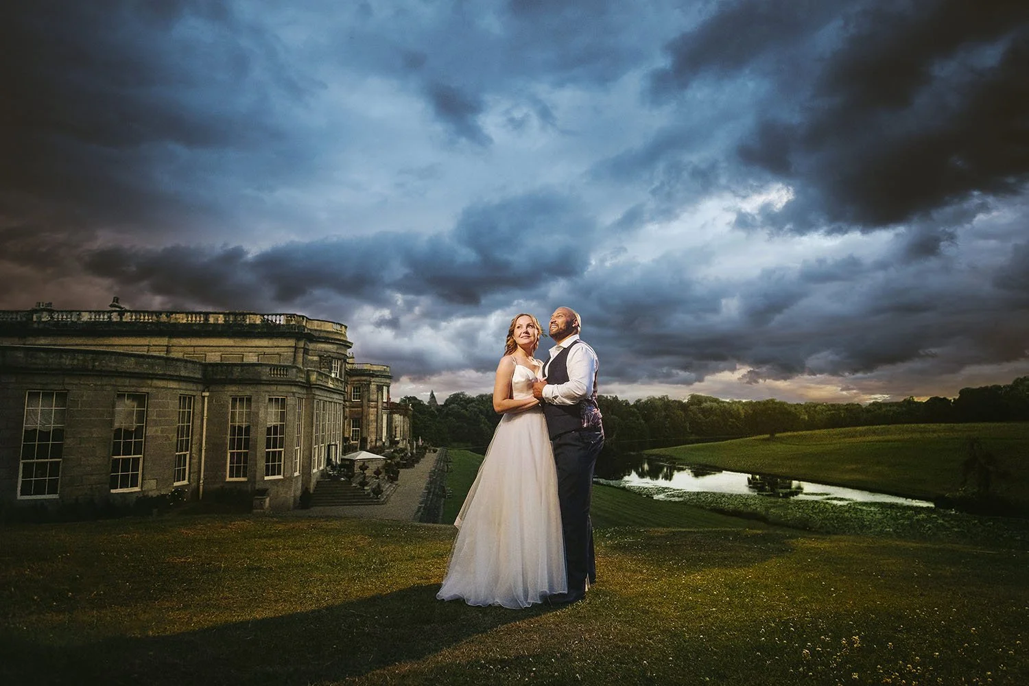 Wynyard Hall wedding: bride and groom embracing on a lawn in front of a stately home beneath a stormy blue sky.