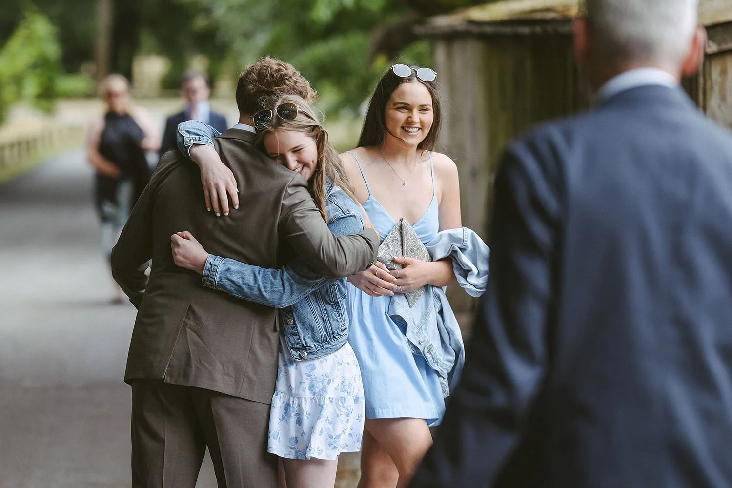The Fig House wedding: wedding guests hugging on a tree-lined path, a young woman in a denim jacket smiling over a man's shoulder.