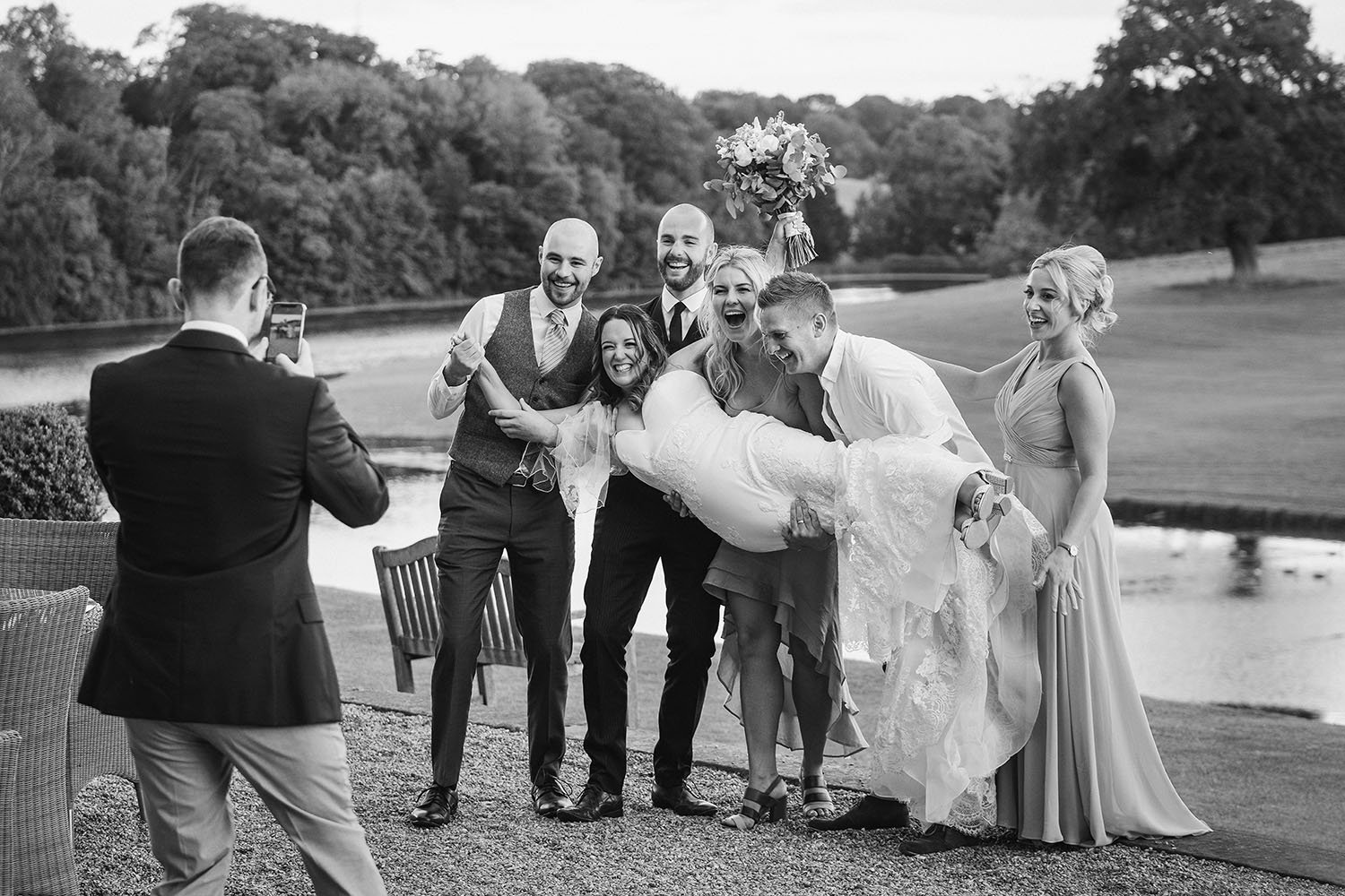 Wynyard Hall wedding: groomsmen playfully lifting the laughing bride beside a lake, in black and white.