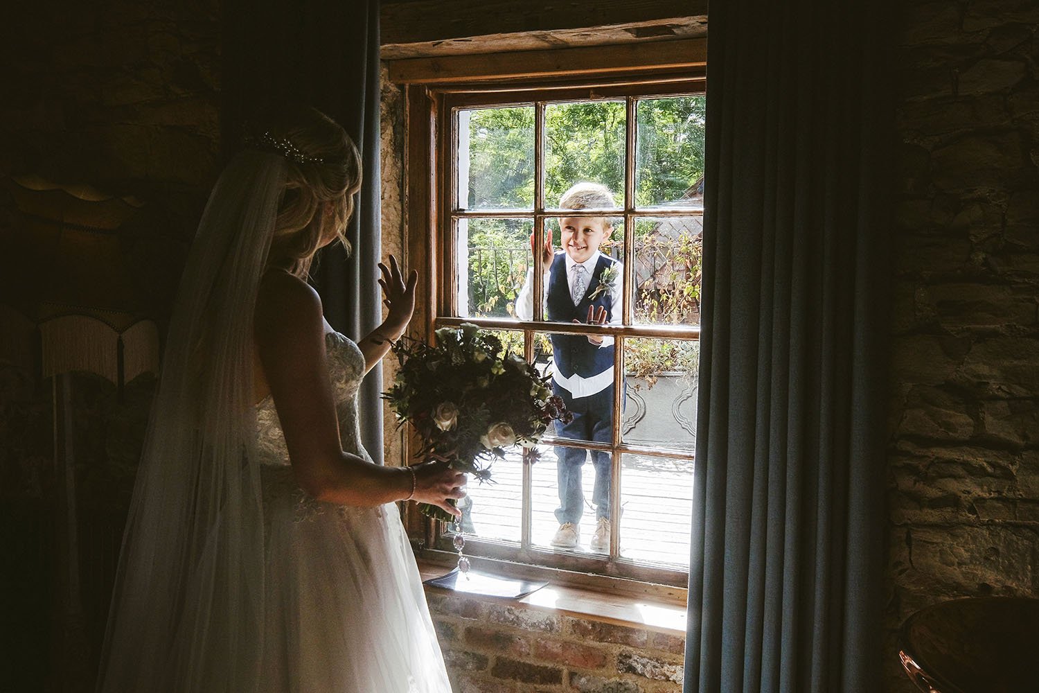 Le Petit Chateau wedding: bride in her veil holding a bouquet at a window as a page boy in a waistcoat peeks in from outside