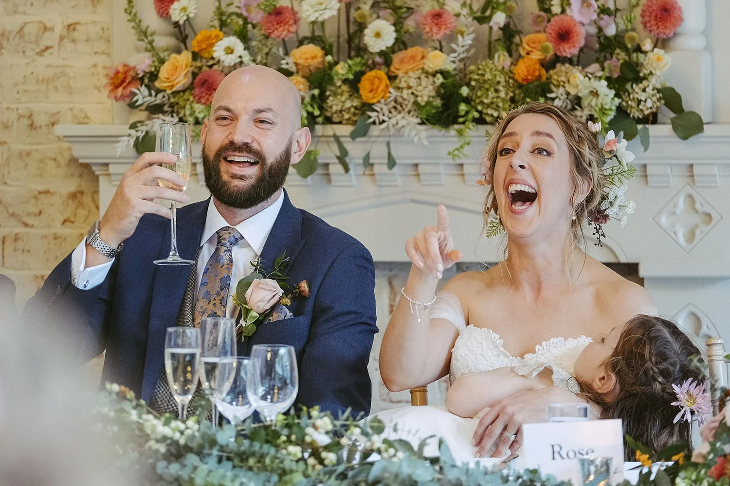 Shotton Grange wedding: bride and groom laughing at the top table with a child on her lap, flowers above the fireplace.