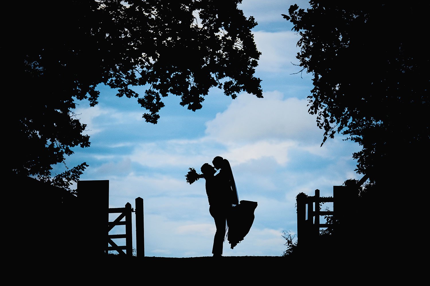 Brinkburn Priory wedding: groom lifting the bride in silhouette between trees and a gate against a blue sky