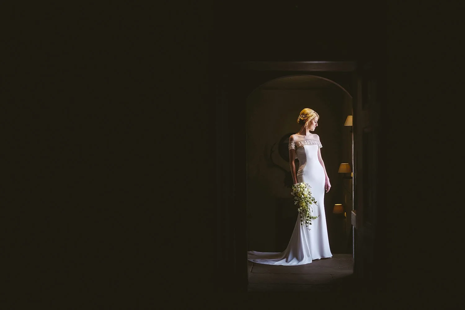 Healey Barn wedding: bride standing with her bouquet in a dark doorway lit by dramatic window light