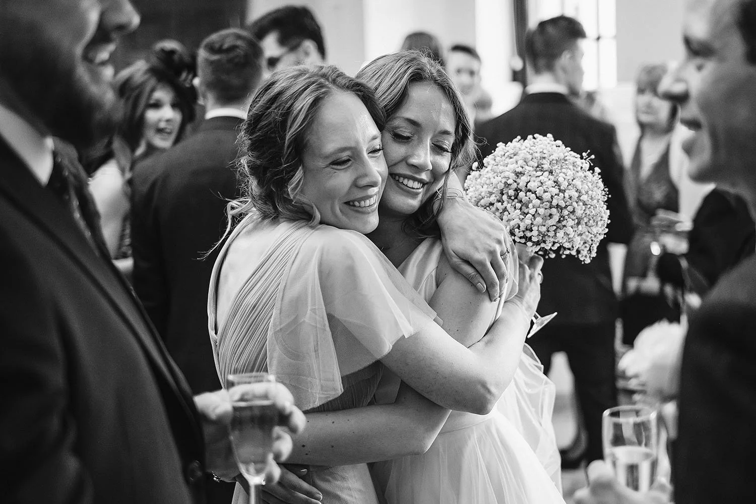Black-and-white wedding reception photo of two smiling women embracing, with guests and a bouquet of baby's breath flowers in the background.