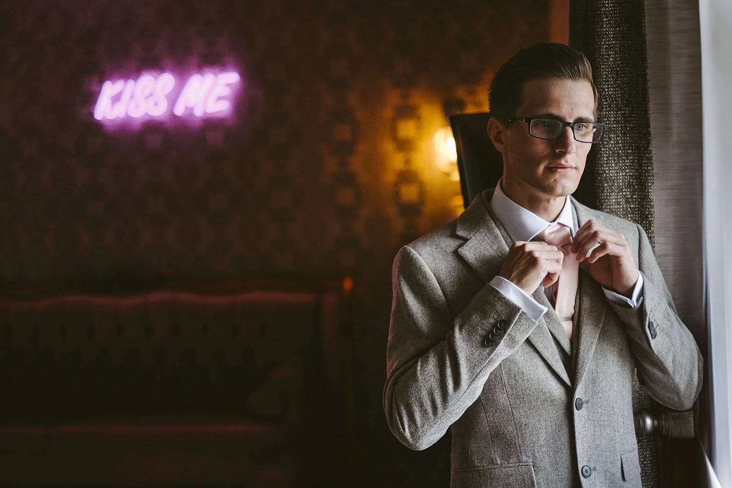 Charlton Hall wedding: groom in glasses and a grey jacket adjusting his tie by a window in a dimly lit bar.