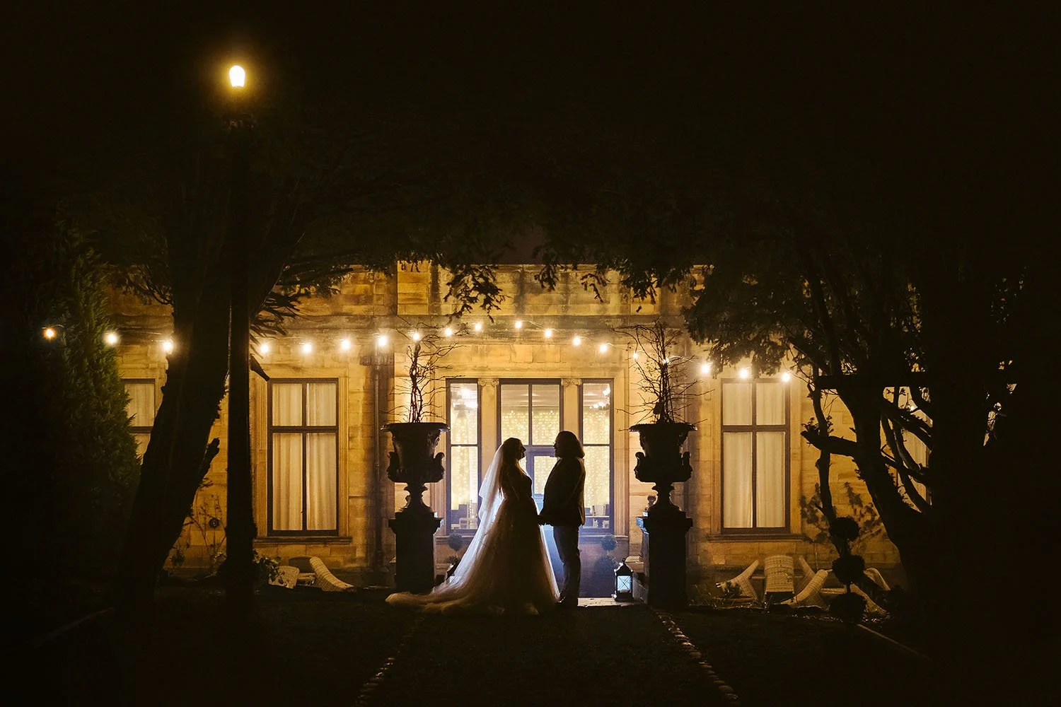 Beamish Hall wedding: bride and groom in silhouette at night in front of the lit stone house, with festoon lights and urns.