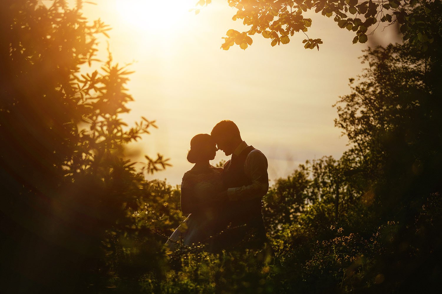Bride and groom in silhouette with foreheads touching against a golden sunset through the trees