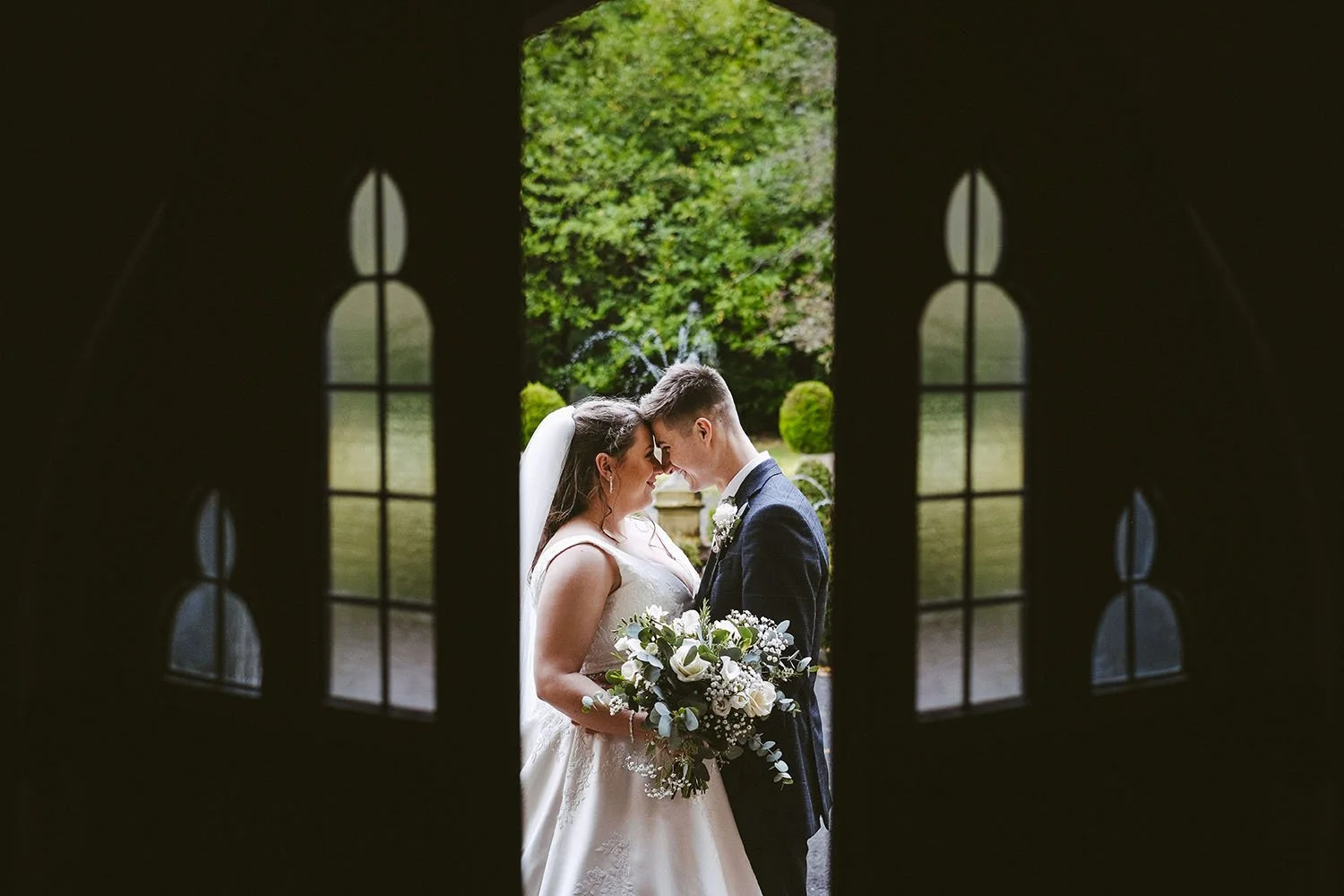 Bride and groom touching foreheads in a dark doorway framed by gothic windows.