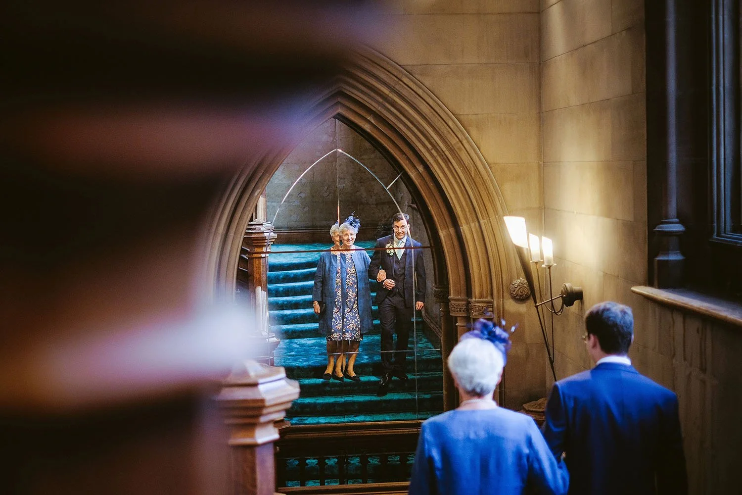 Matfen Hall wedding: mother of the groom and the groom walking down a staircase seen through a gothic archway
