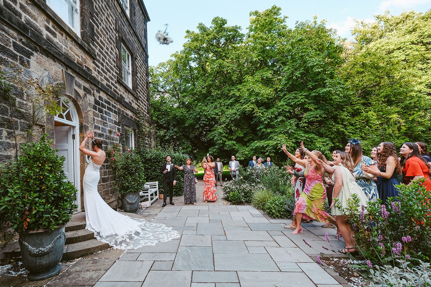 Bride throwing her bouquet over her shoulder as guests reach up on a stone patio beside a flower garden.