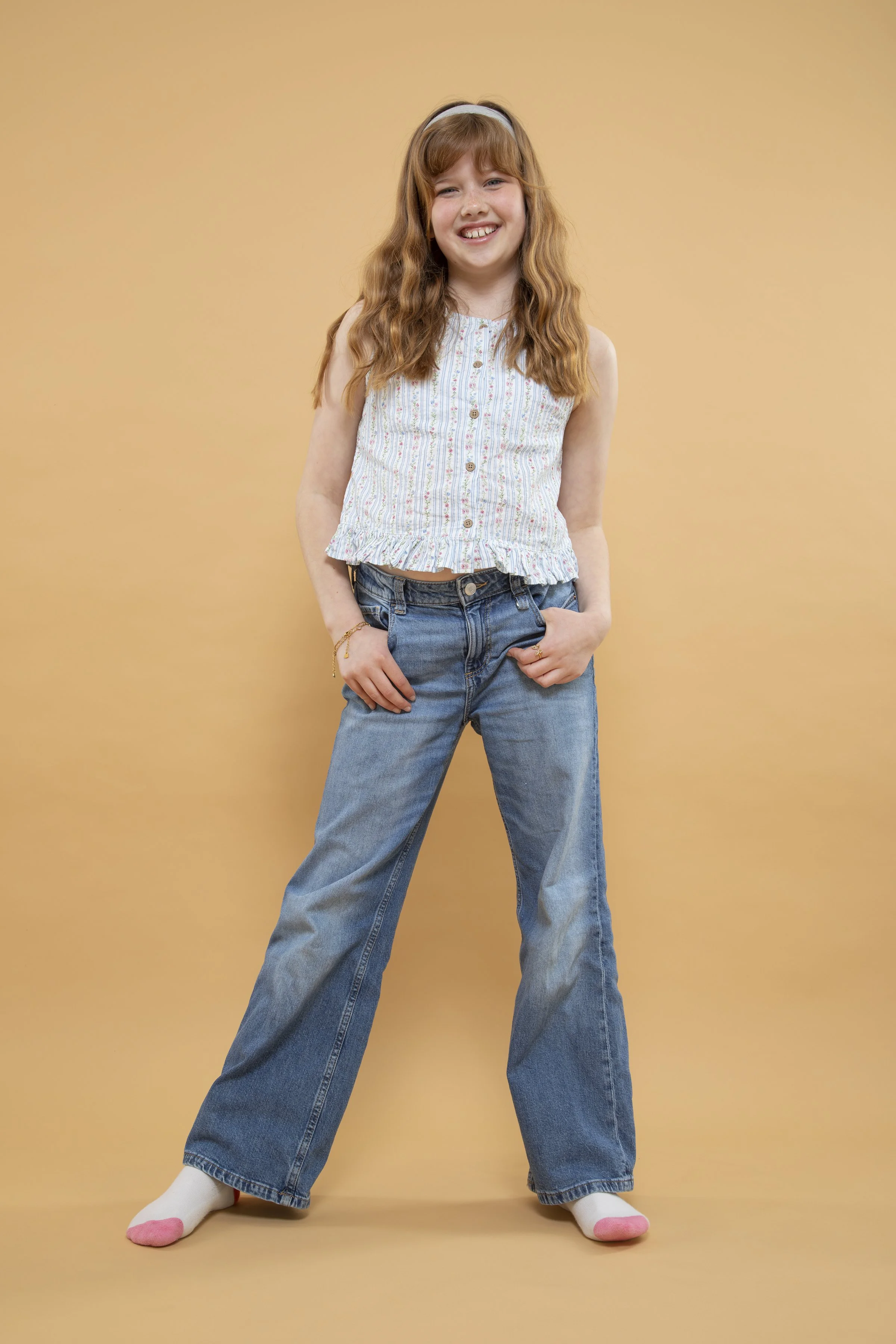 A young girl with long, wavy red hair, wearing a sleeveless, buttoned, striped top, baggy jeans, and white socks with pink toes, standing against a plain tan background, smiling at the camera.