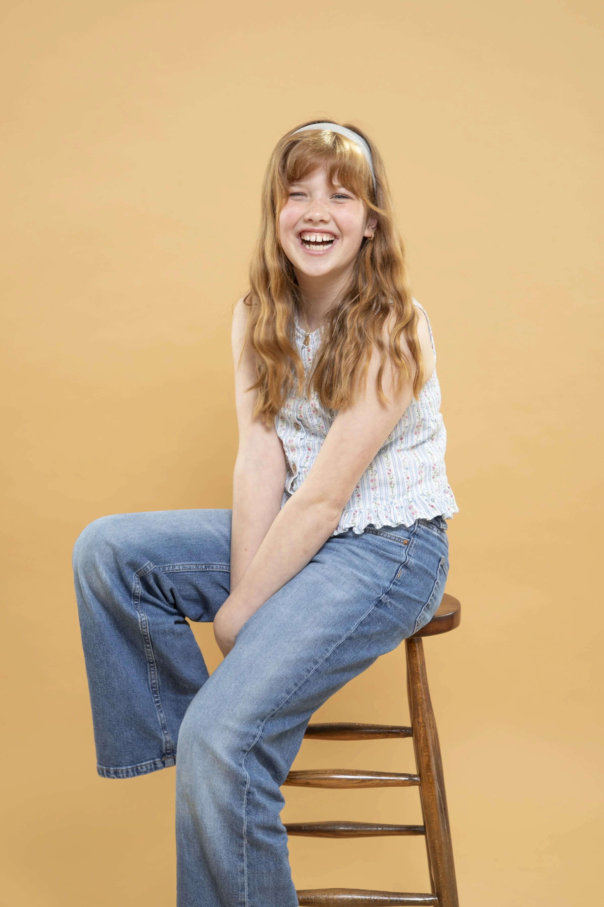 A young girl with long red hair, wearing a sleeveless striped top and blue jeans, sitting on a wooden stool against a yellow background, smiling and laughing, photographed by Garter Wedding Photography.
