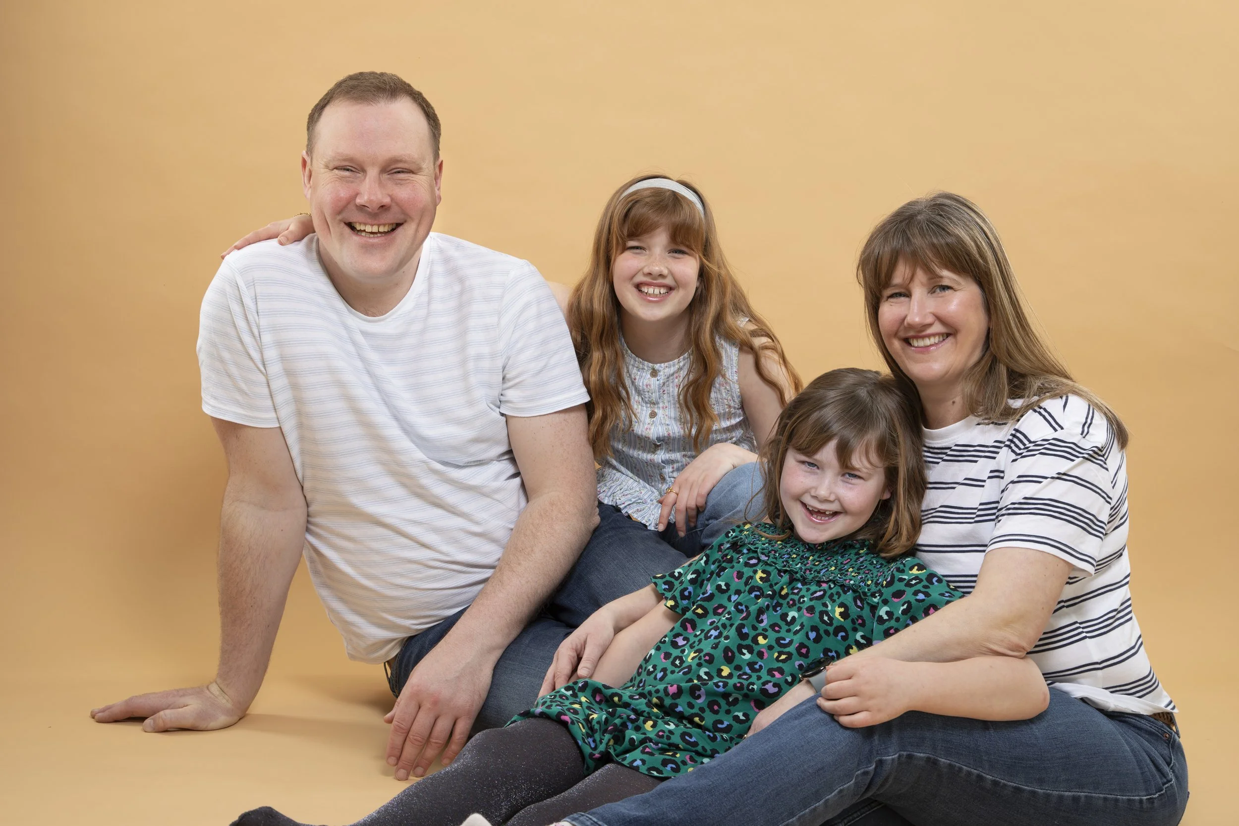 A happy family of four sitting and lying on a yellow background. The father and mother are smiling, with the father wearing a white T-shirt and the mother in a striped shirt. Their two daughters, with brown hair and wearing colorful dresses, are also