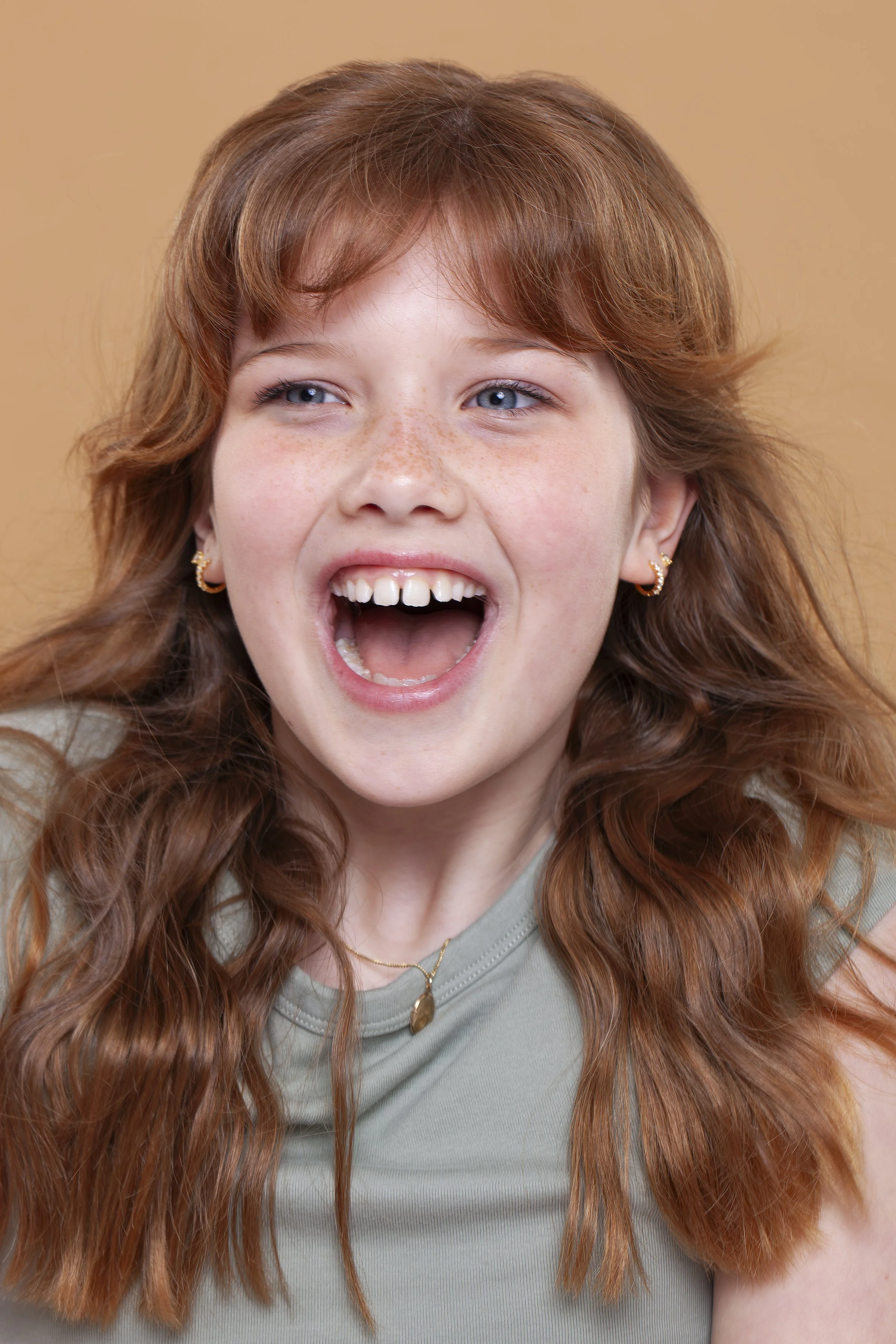 Close-up portrait of a young girl with red hair, blue eyes, freckles, smiling with her mouth open, wearing gold earrings and a necklace, against a beige background photographed by Garter Wedding Photography.
