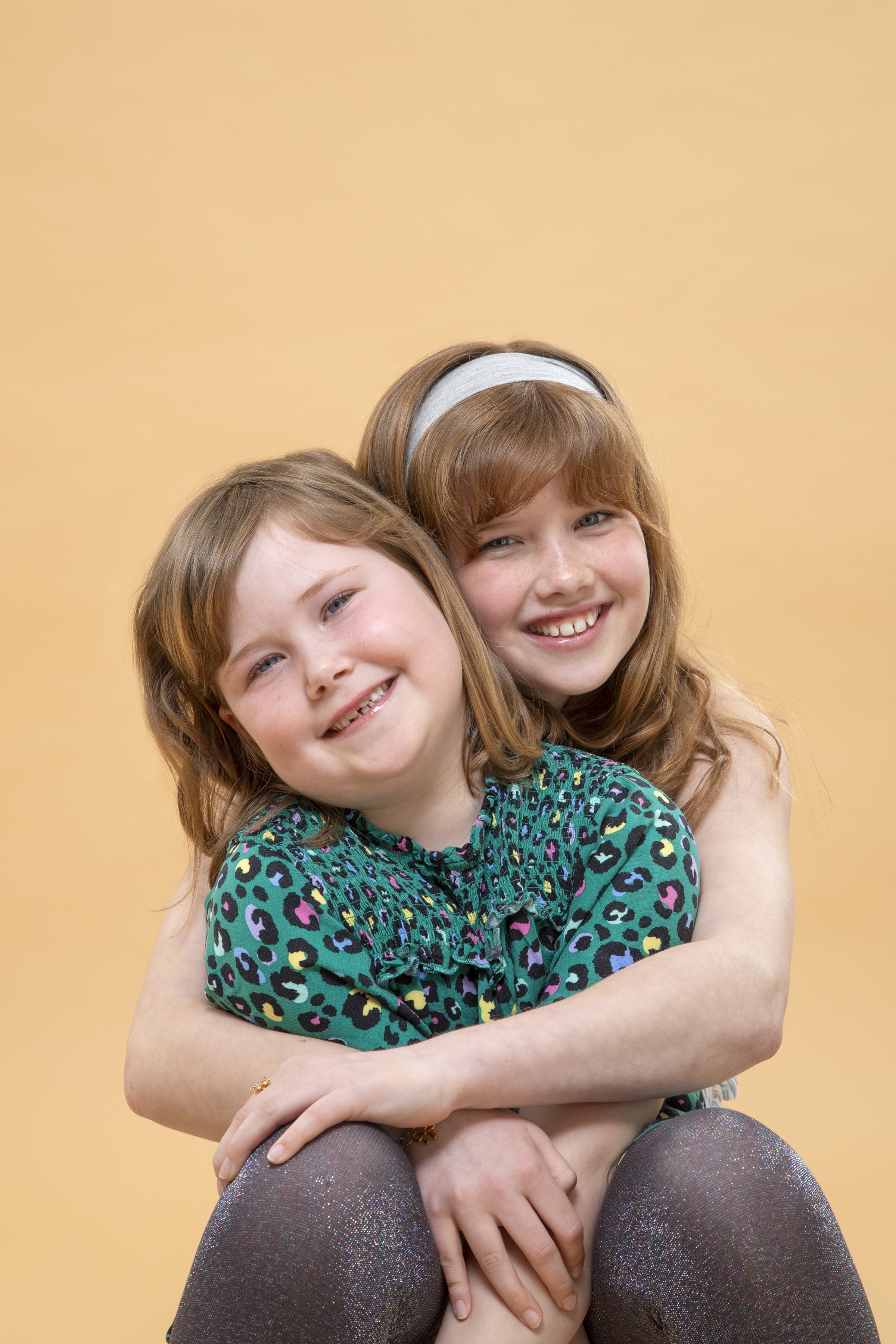 Two young girls with red hair smiling and embracing each other against a solid beige background.