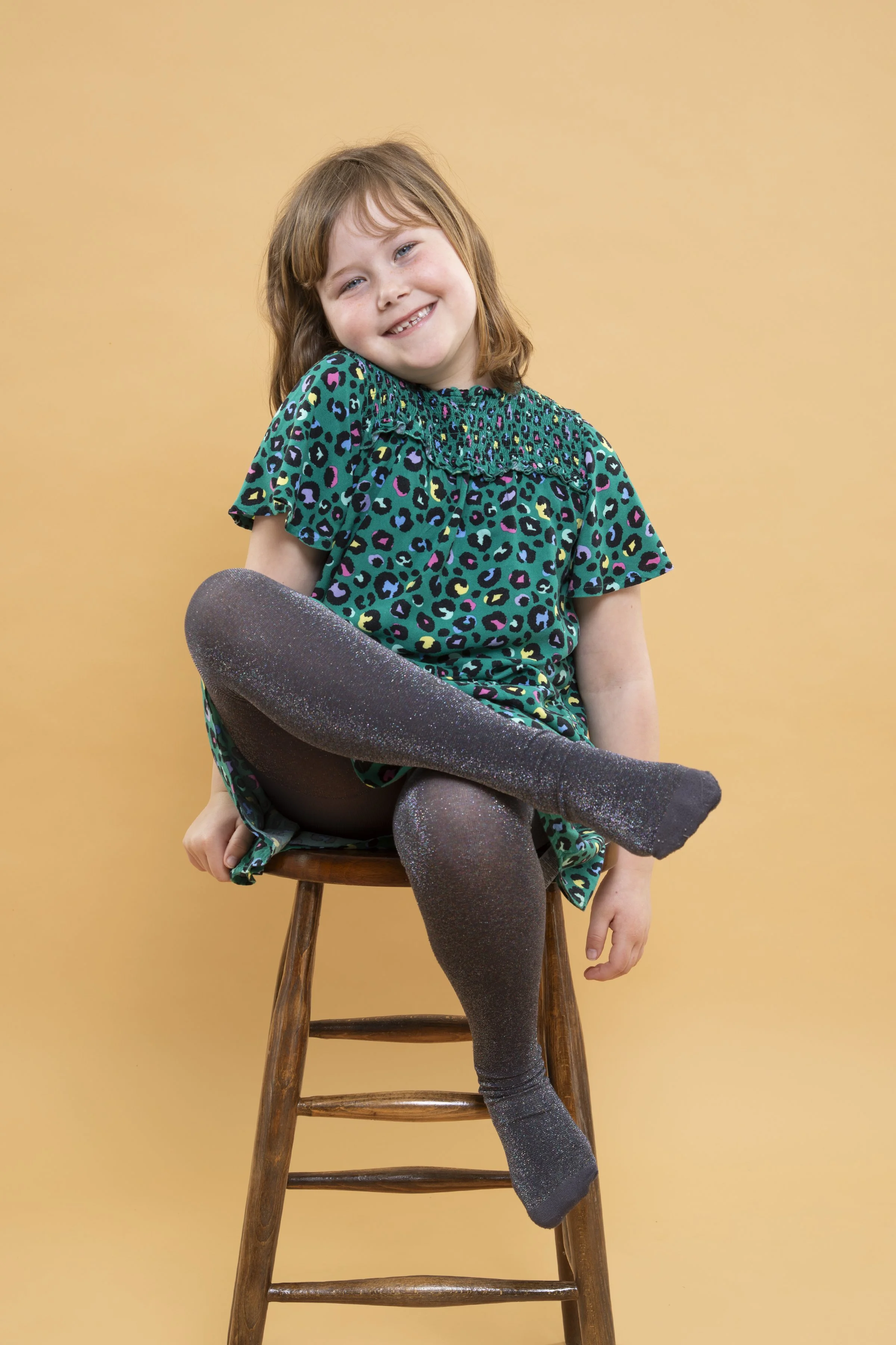 A young girl sitting on a wooden stool with one leg crossed over the other, smiling at the camera, wearing a colorful leopard print dress and glittery black tights, against a plain light orange background.