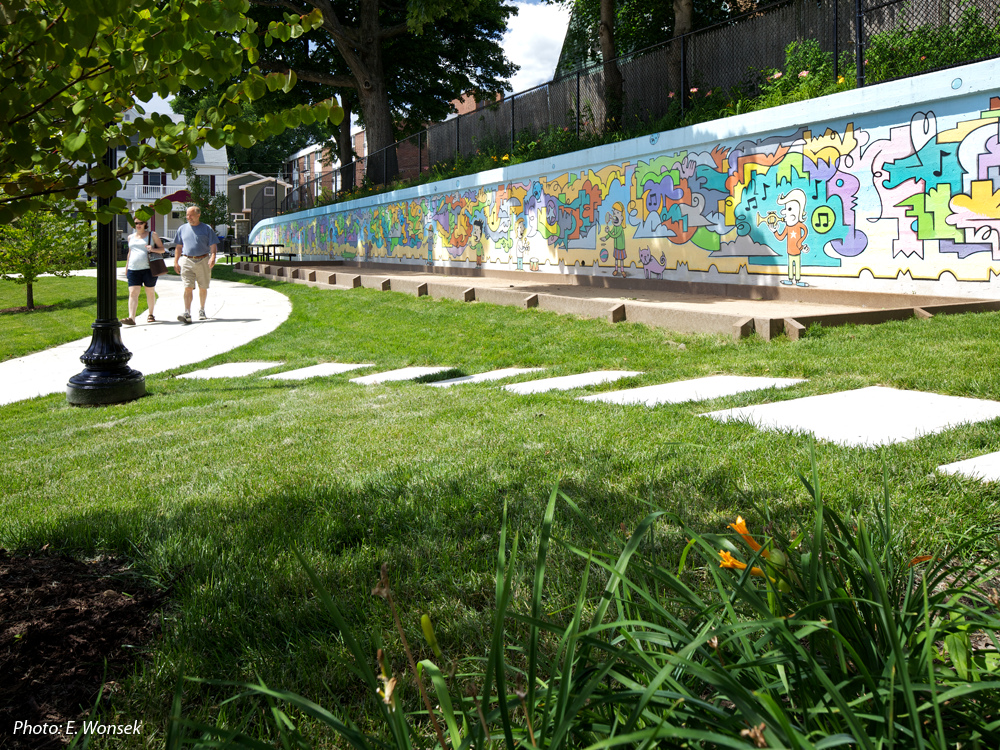 Formerly separated onto two levels, this park has been re-connected through our plan. An accessible route now links the refurbished play area and seating on the lower half, with a revitalized upper level that replaced an unusable basket-ball court with open lawn, a bocce court, picnic tables, and a winding pathway. Inspired by a recent mural on the uppermost wall, CBA's design uses the same vibrant colors, echoes similar shapes, and draws out details such as a blue bear's face to form the basis of the spray area.