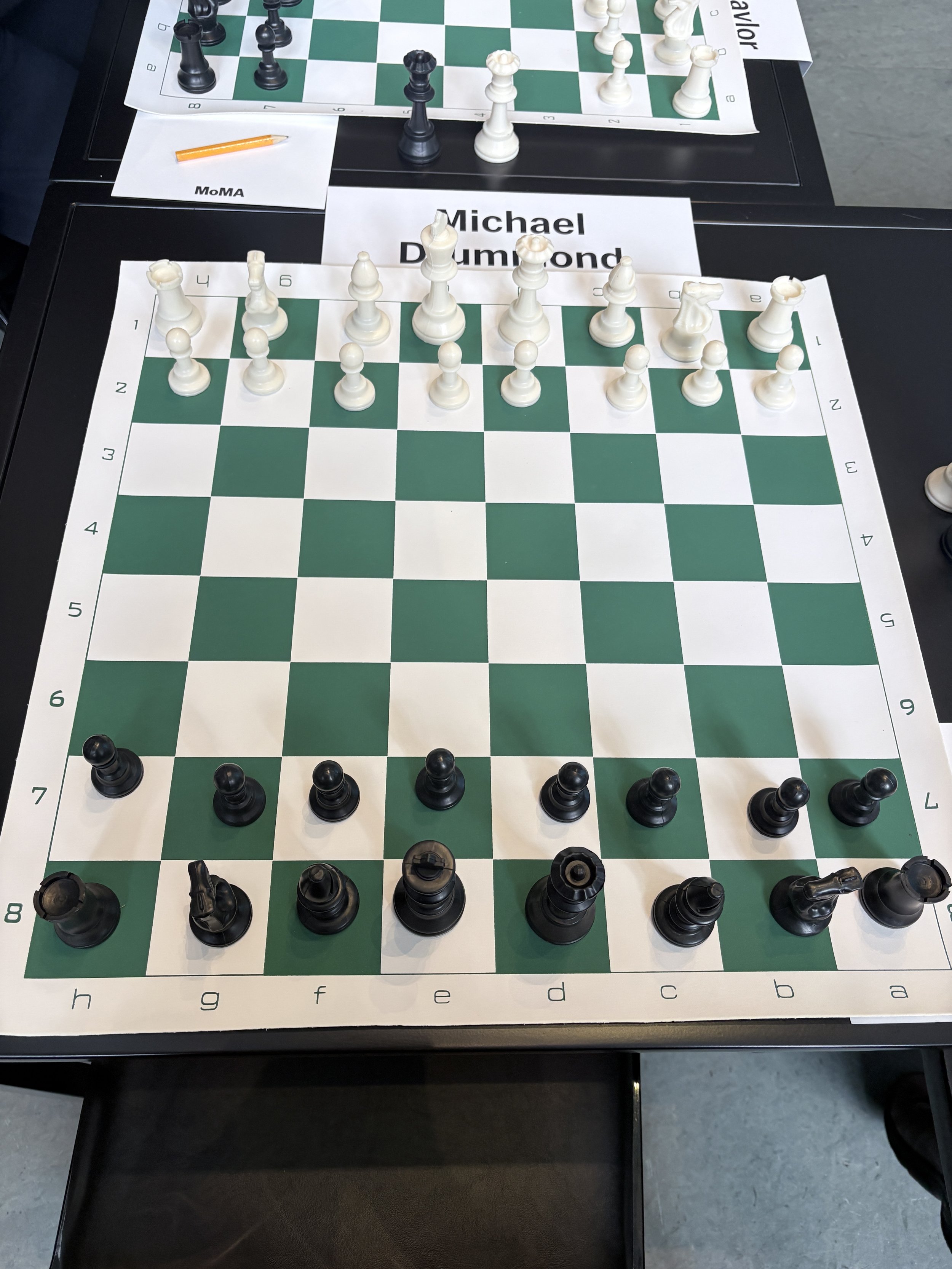 a green and white chessboard with black and white chess pieces are set at the beginning of a game with the name Michael Drummond placed in front