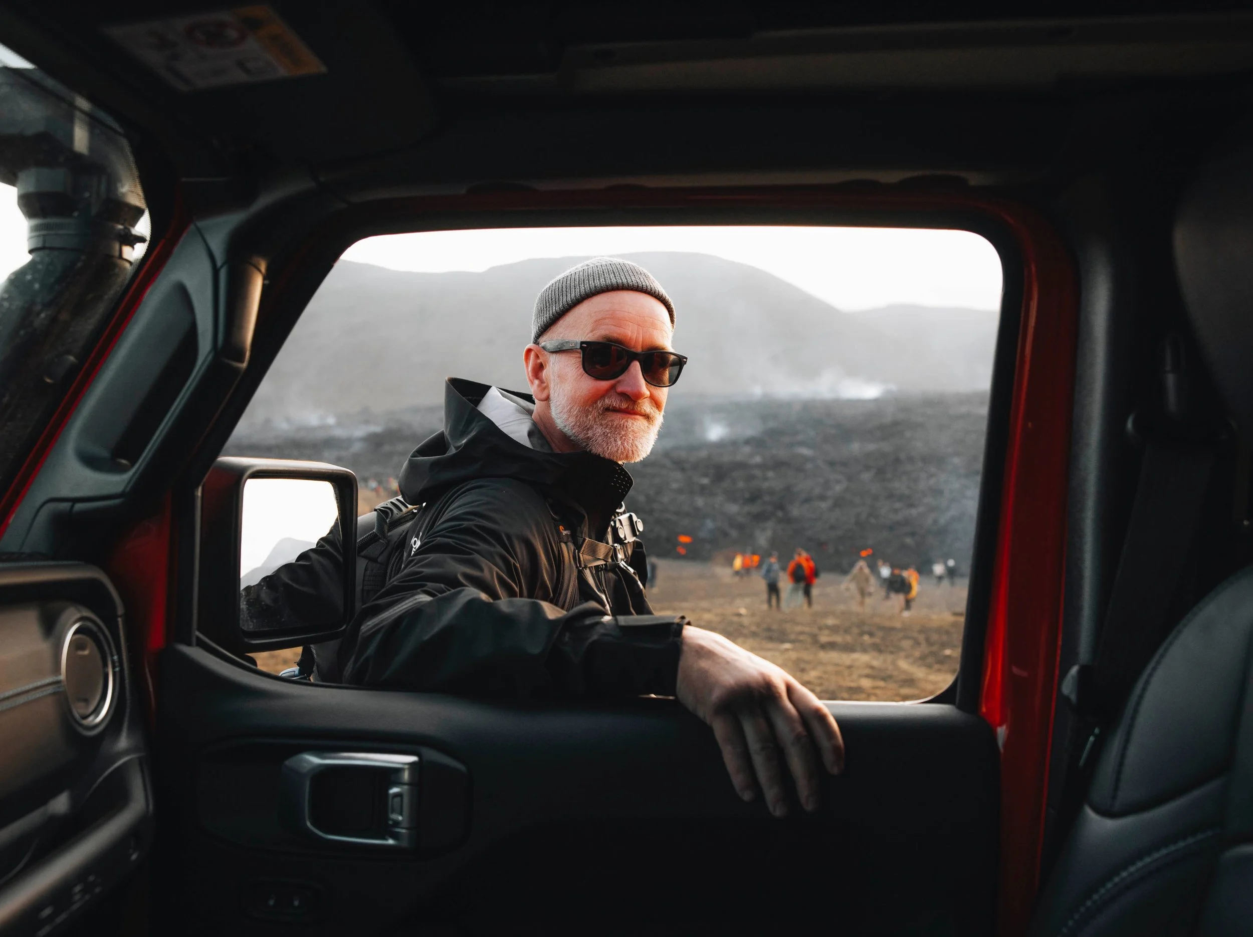 Siggeir wearing sunglasses and a gray beanie looking out the window of a red vehicle with a volcano and cloudy sky background.