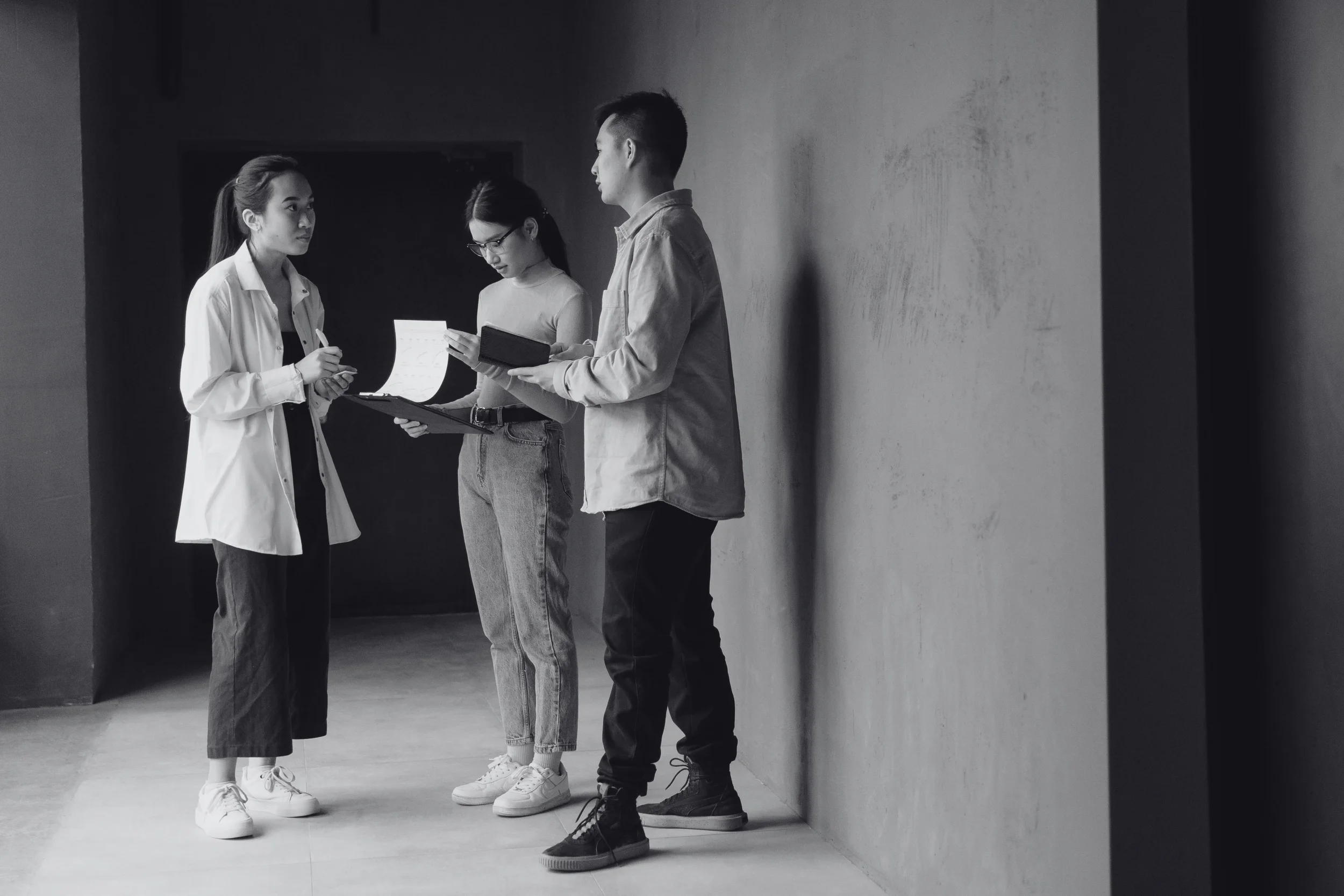Three young adults standing in a hallway, engaged in conversation and reviewing documents.