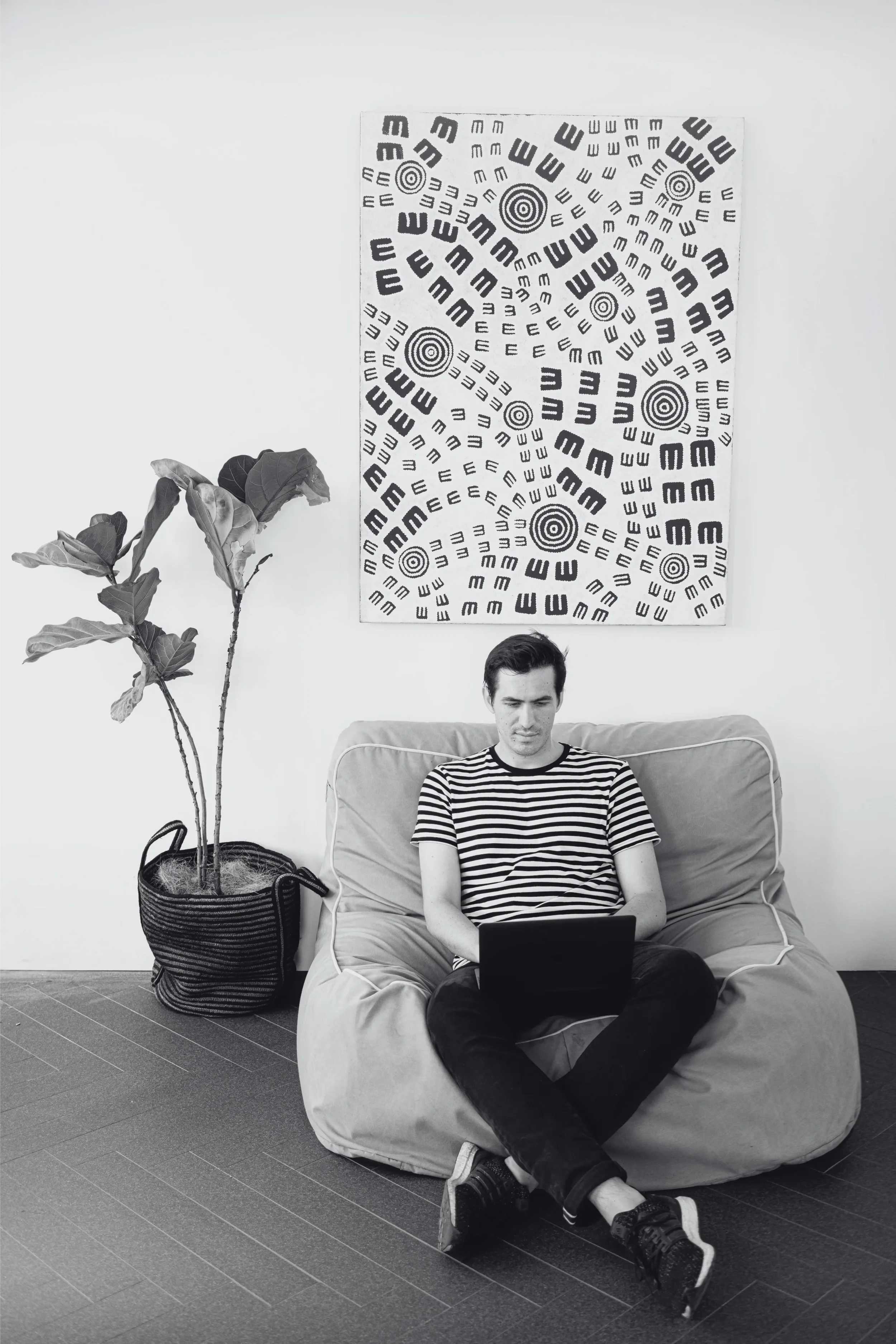 A man sitting on a beanbag chair, looking at a tablet. Behind him is an abstract black and white artwork on the wall. To his left is a potted plant with large leaves in a striped basket.
