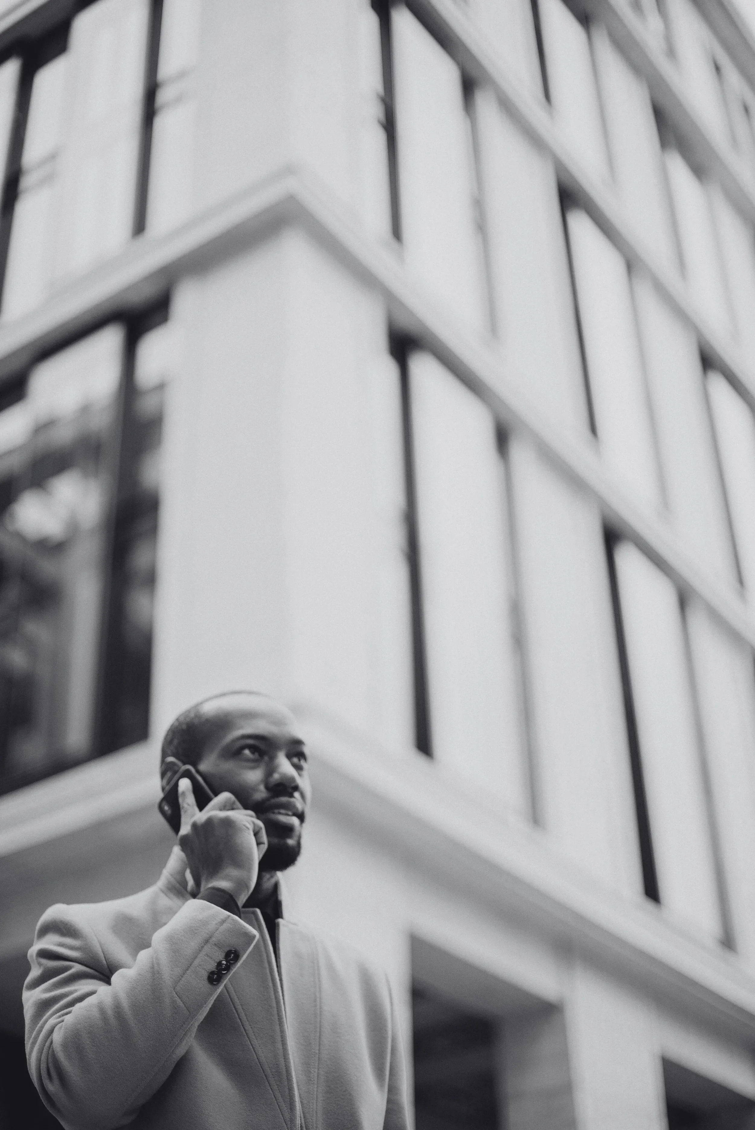 A man in a light-colored coat talking on a cellphone outdoors in front of a modern glass office building.
