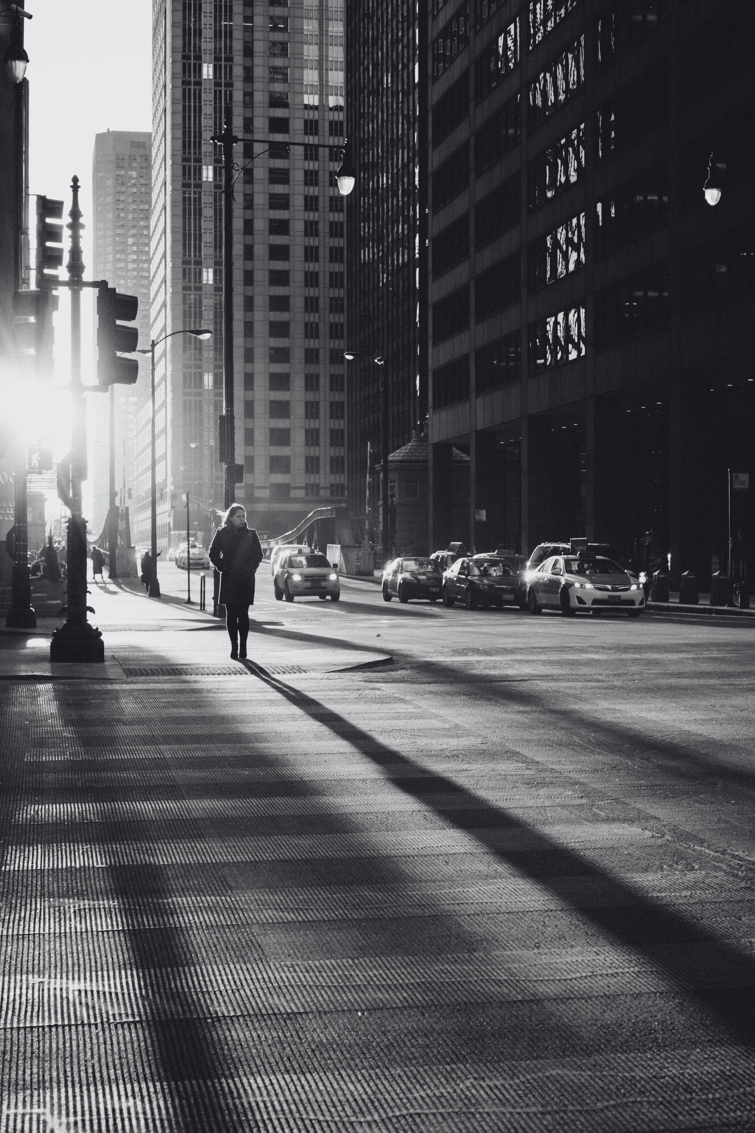 A black and white photo of a city street with a woman walking on the sidewalk and cars parked along the curb, sunlight casting long shadows.