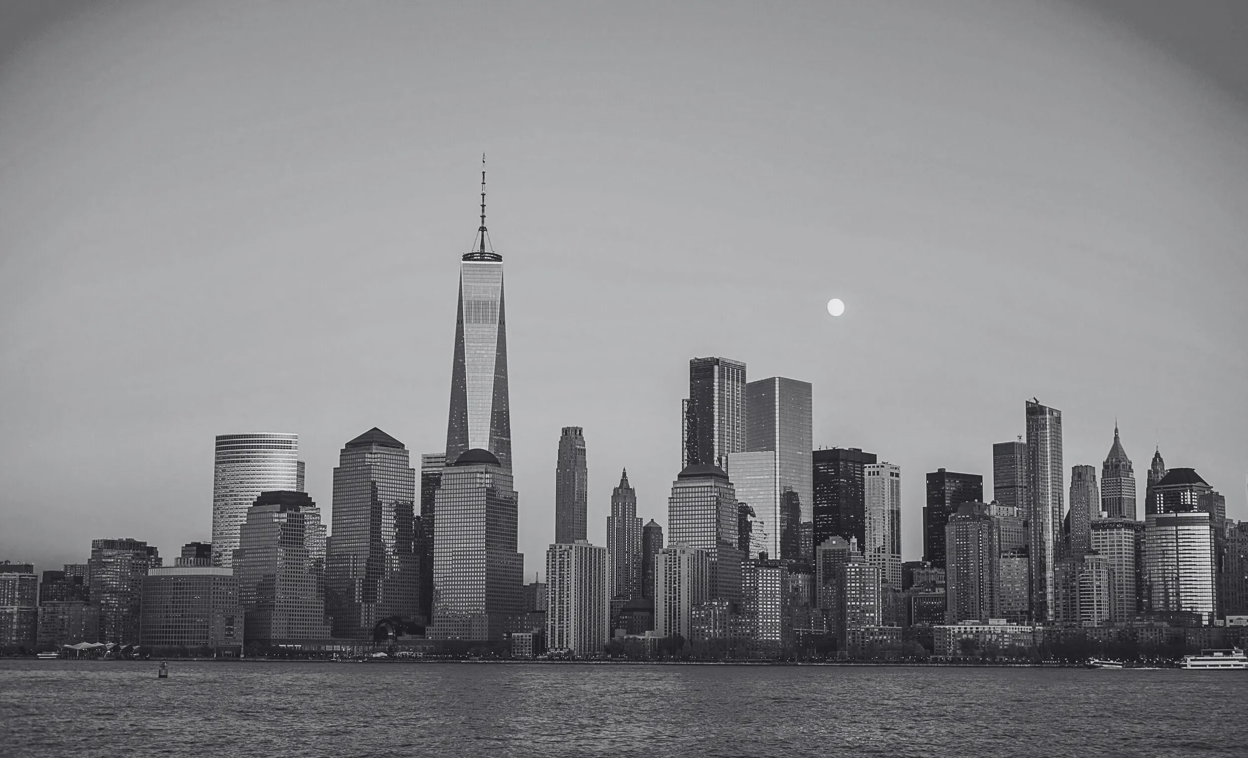 Black and white photo of the New York City skyline with one World Trade Center building, skyscrapers, and a full moon over the water.