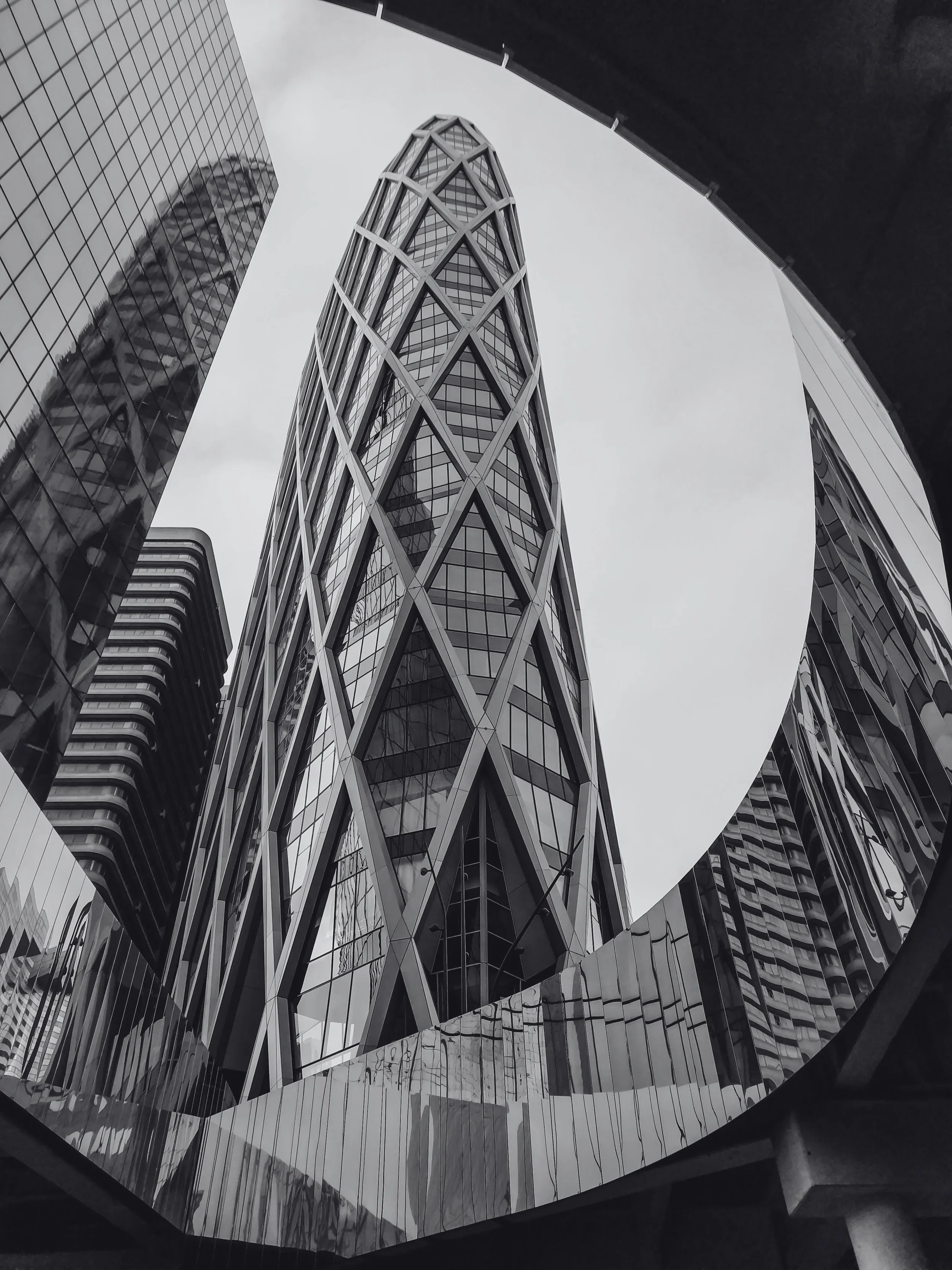 A black and white photo of a modern skyscraper with a glass facade, taken from below through a circular frame, with other high-rise buildings around it.
