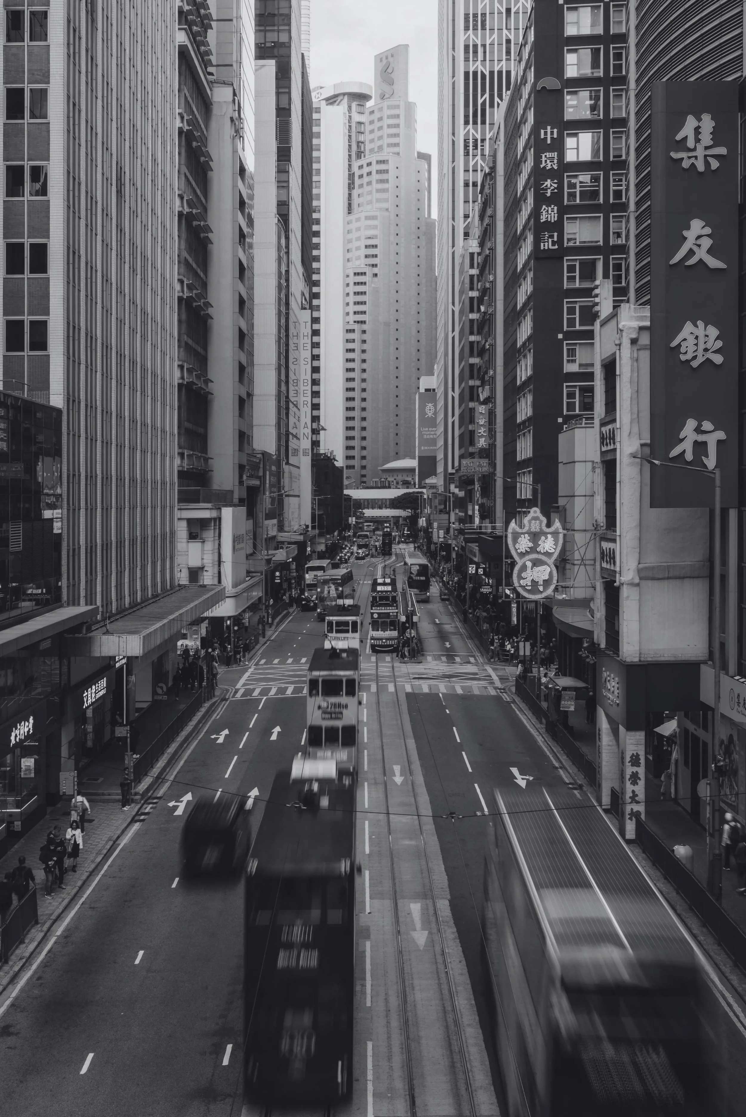 Black and white photo of a busy city street with tall skyscrapers, double-decker buses, and pedestrians.