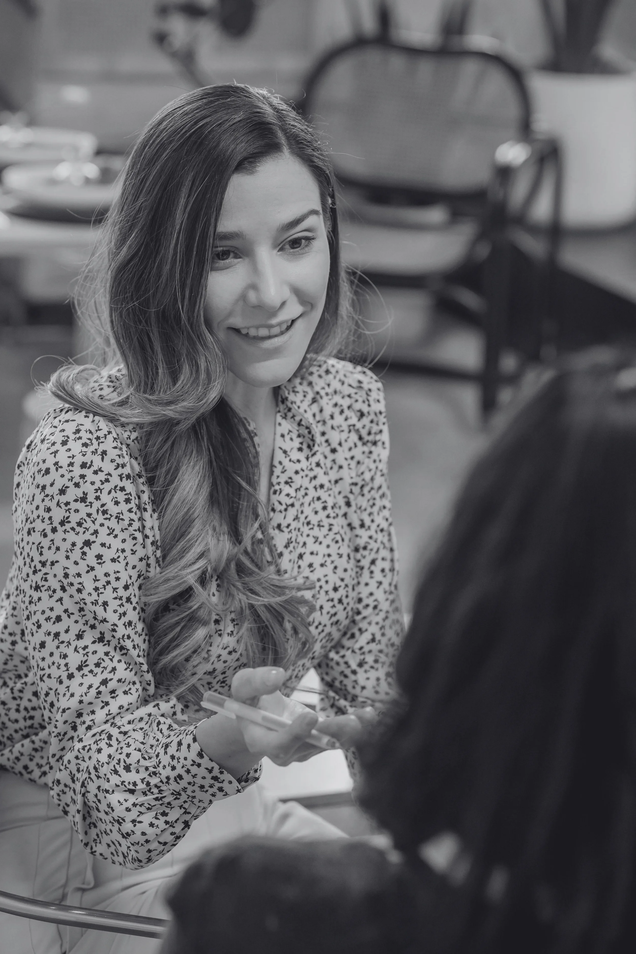 A smiling woman with long wavy hair holding a pen, engaged in a conversation in a café.