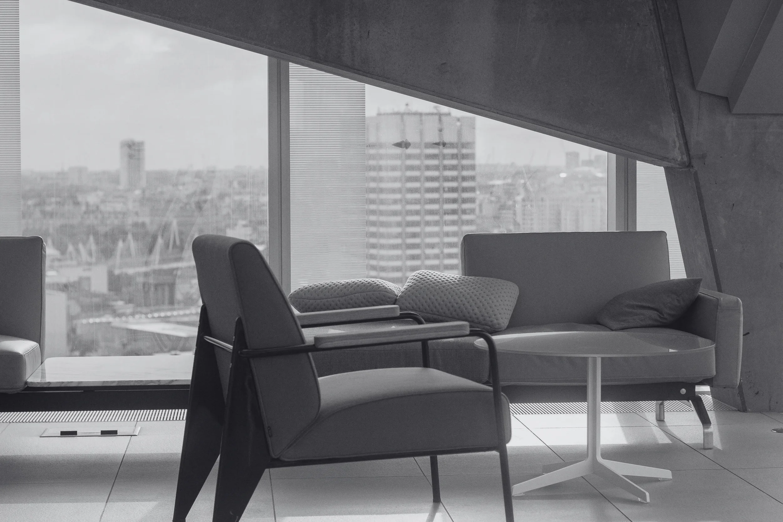 Modern office lounge with chairs and a sofa, large floor-to-ceiling windows showing a cityscape, and an overhanging concrete ceiling
