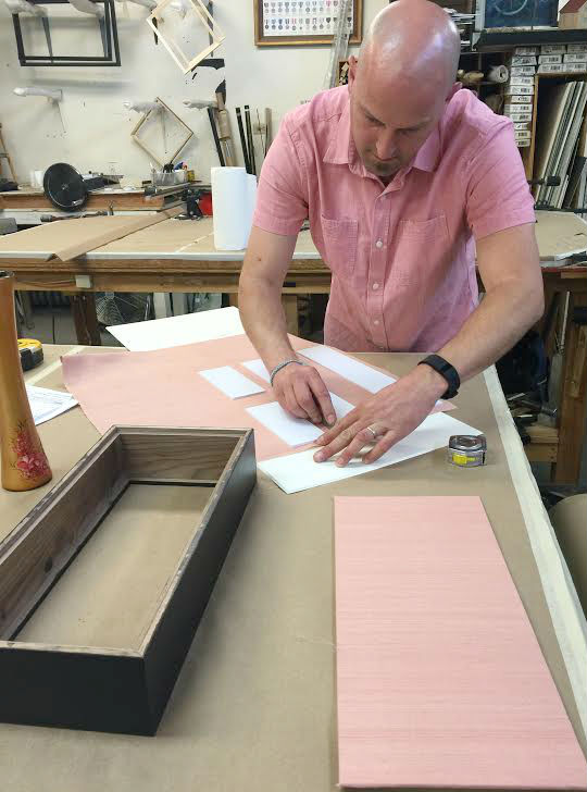 Our framer/owner Eric, mapped out the lining of the shadow box walls. Each side ishand-wrapped with a pink, silk fabric. Eric also, and unintentionally, matched the light pink silk fabric. 
