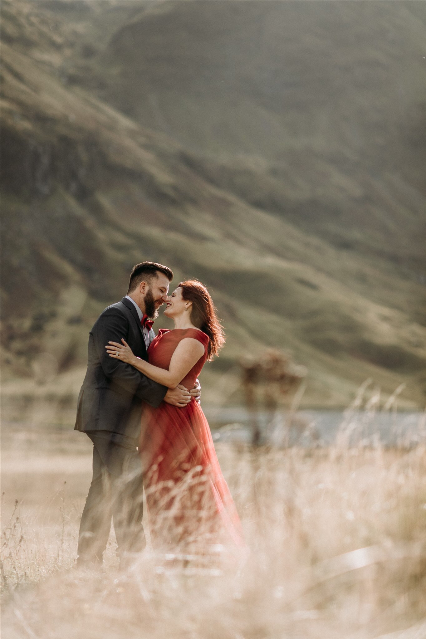 Glen Coe Scotland elopement. Bride in red dress and her groom holding each other and smiling in a field in the Scottish Highlands | Adventure elopement photographer