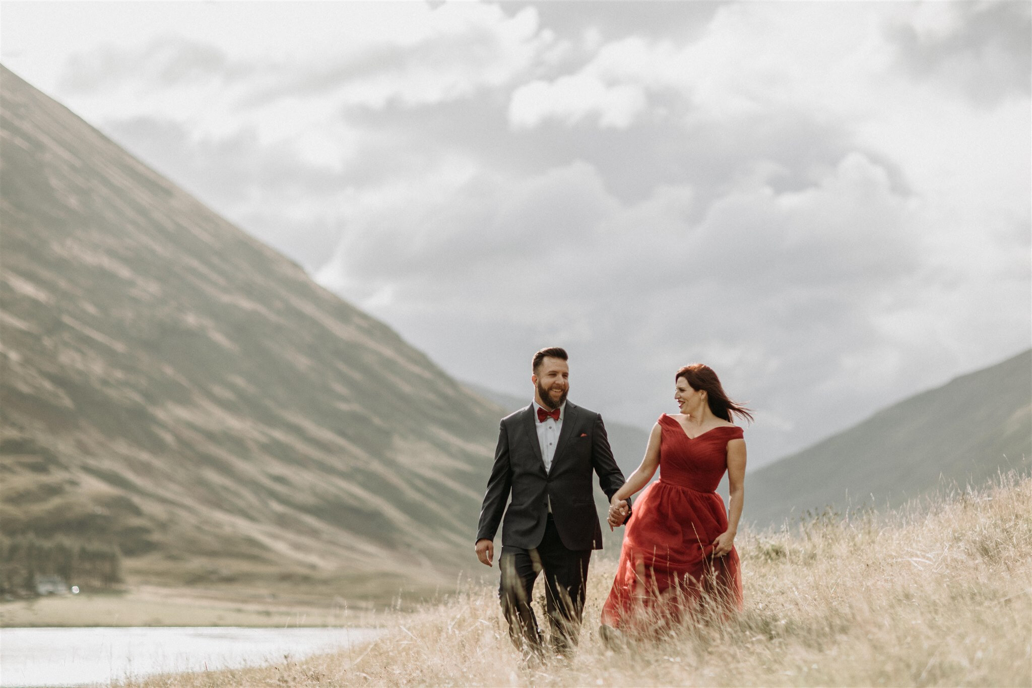  Glen Coe Scotland elopement adventure session. Bride in red dress and her groom walking hand in hand through a field in the Scottish Highlands | Adventure elopement photographer