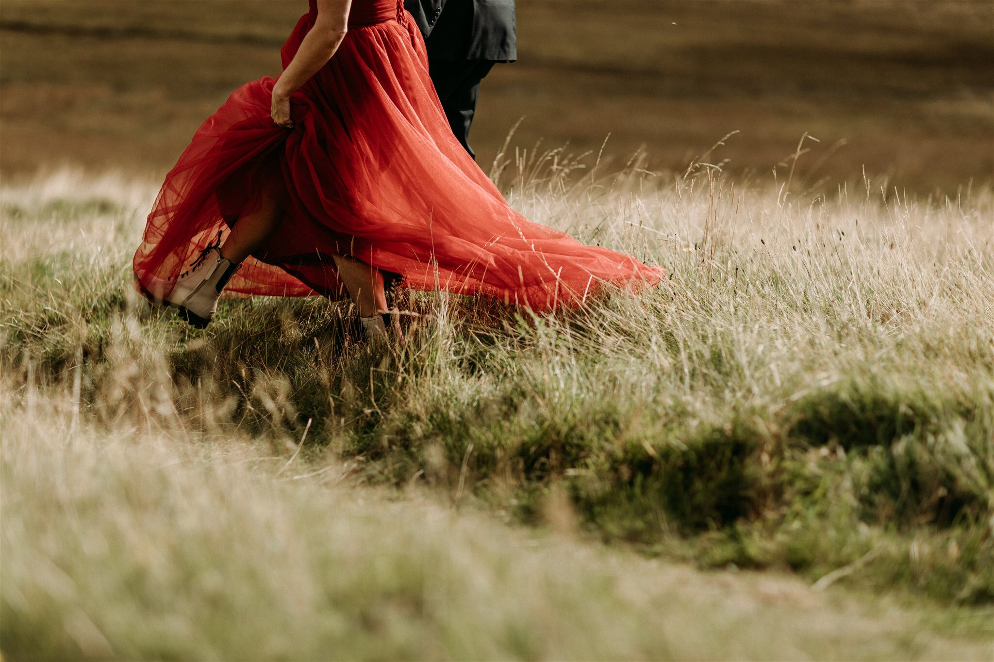 Glen Coe Scotland elopement adventure session. Bride’s red dress blows in the wind as she and her groom walk through a field in the Scottish Highlands | Adventure elopement photographer