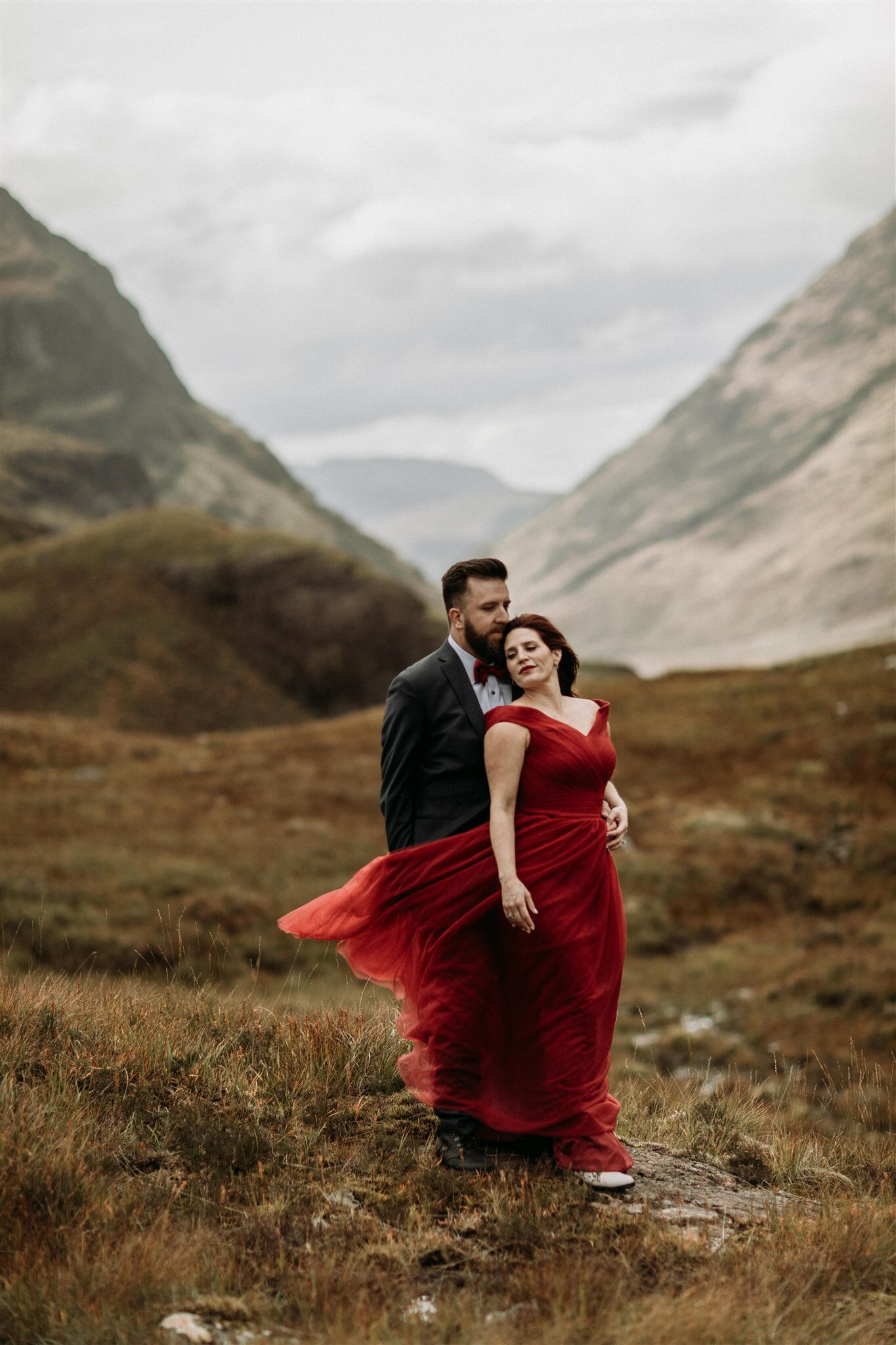Glen Coe Scotland adventure elopement. Bride in red dress blowing in the wind stands with her back to her groom’s chest in the Scottish Highlands | Adventure elopement photographer