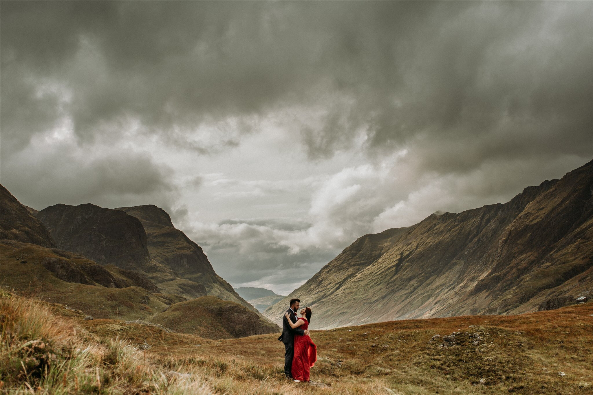 Glen Coe Scotland elopement adventure session. Bride in red dress and her groom holding each other and smiling in a field in the Scottish Highlands | Adventure elopement photographer